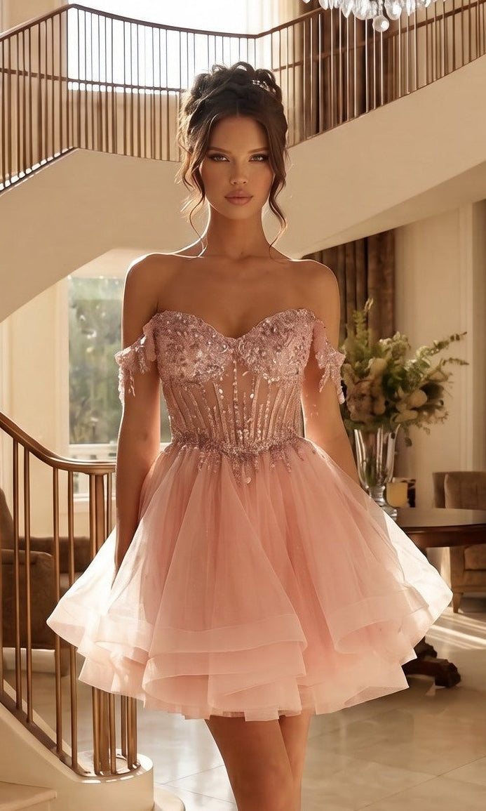 A woman stands indoors wearing the Short Champagne Corset Prom Dress: Juliet JT947L in Rose, a stylish off-the-shoulder tulle dress with floral embellishments, beaded fringe, and a fitted bodice. Flowers and a staircase complete the elegant setting.